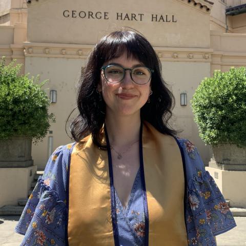 Sarah is standing in the middle of the frame, facing the camera. She is wearing a graduation stole over a lavender floral blouse. Behind her is a building with the letters "George Hart Hall" welded onto the face of the building. 