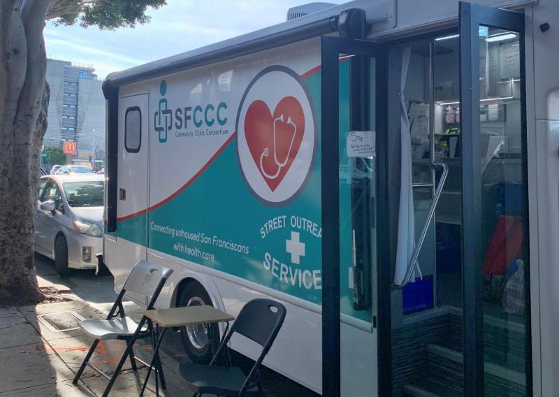 A photo of the Street Outreach Services (SOS) van, parked. There's a table with two folding chairs on either side of it. On the van, the sign reads "Connecting unhoused San Franciscans with health care."