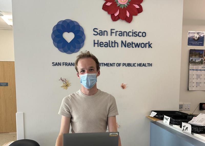John Hansen is standing and facing the camera. Behind them is the San Francisco Health Network logo. 