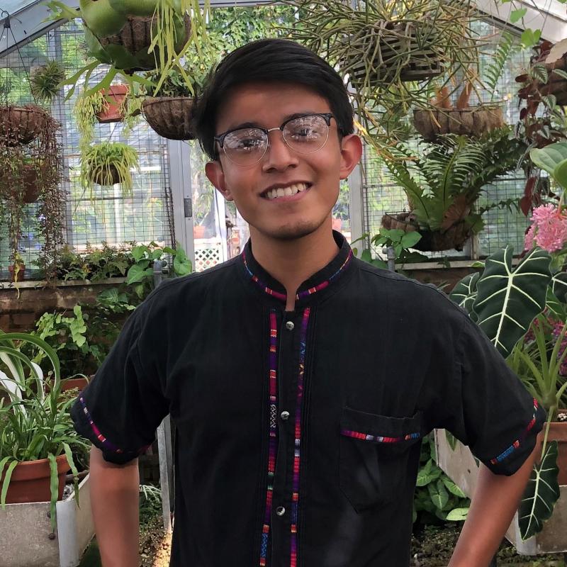 Alex Morales is seen here smiling at the camera, with arms perched on his waist. He is wearing a traditional guayabera with embroidery, and is seen with a background surrounded by hanging plants and potted plants.