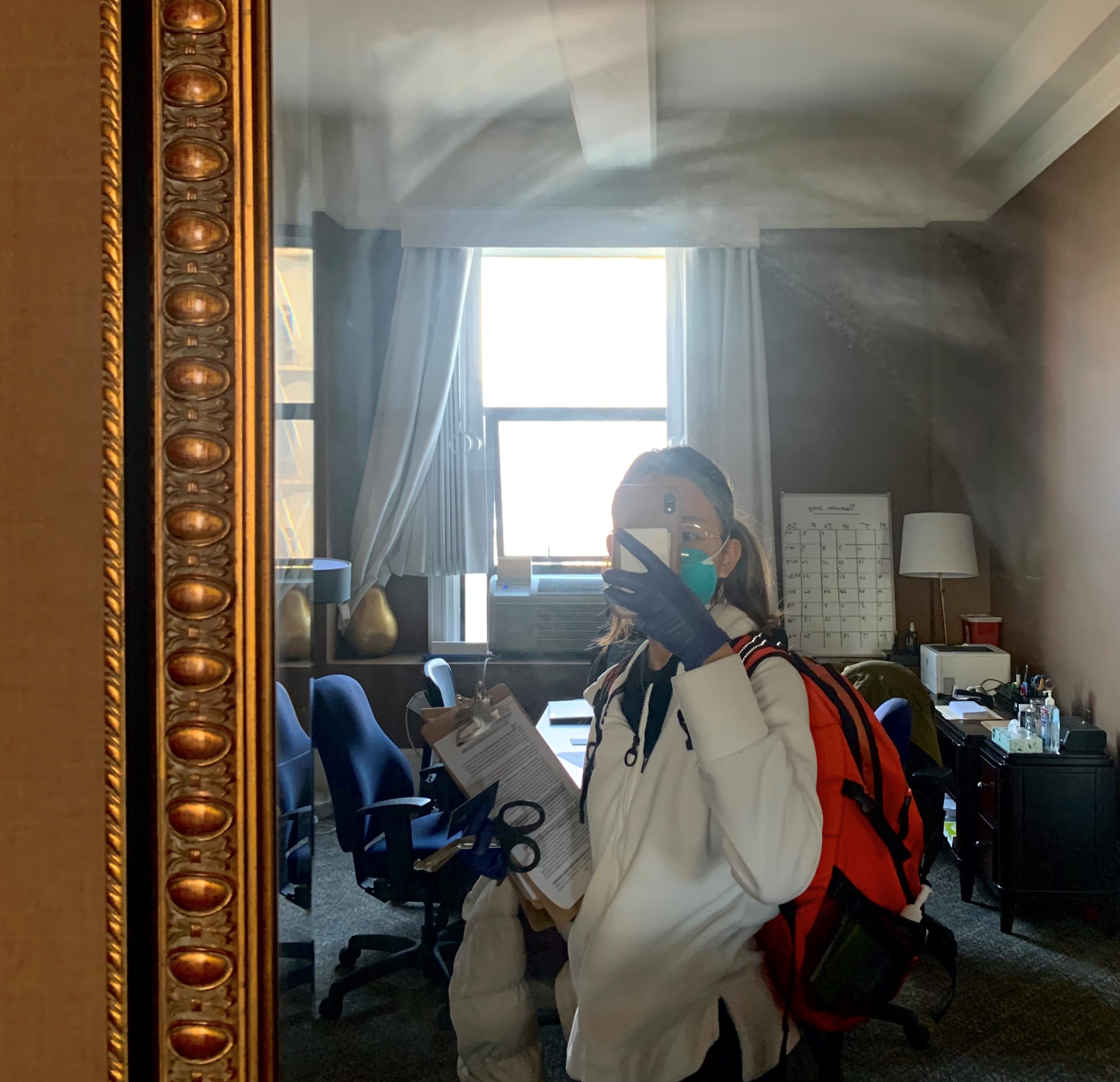 Fern takes a selfie in the mirror at one of the DPH offices. She is holding a clipboard, wearing latex gloves, and is carrying an orange backpack with medical supplies.