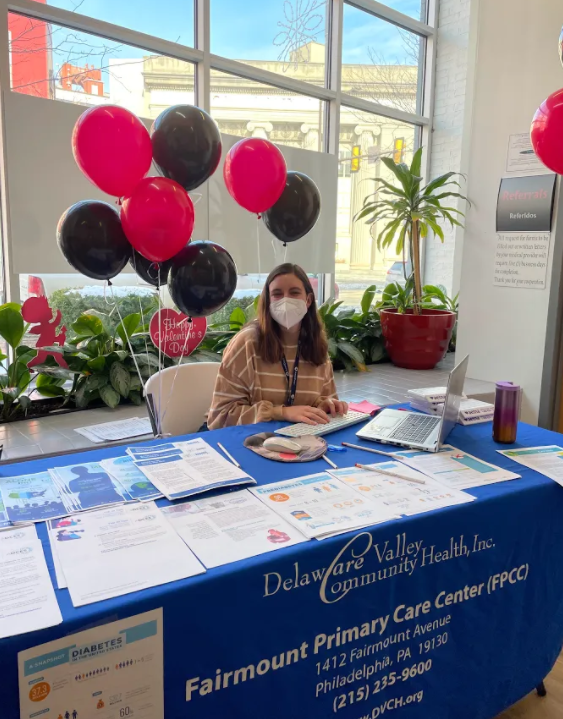 Anne sitting at a table with a blue table cloth that says Fairmount Primary Care Center. On the table are pamphlets containing health information. There are red balloons behind her.