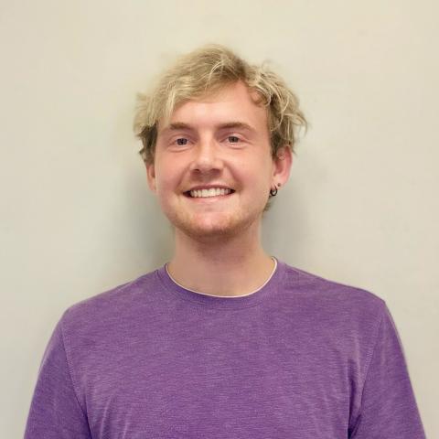Will Crews is facing towards the camera, smiling. He is wearing a purple t-shirt and is against a white, blank wall.