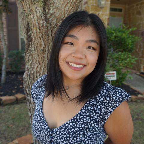 Angela Zhang stands in the middle of the frame, her arms behind her back, smiling at the camera. She is wearing a navy dress with white floral patterns. Behind her is a tree.