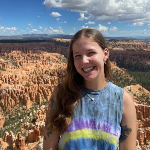 Grace Lemke is standing to the middle right of the frame, smiling at the camera. She is wearing a tie-dyed tank top. Behind her is a canyon with mountains in the far background.
