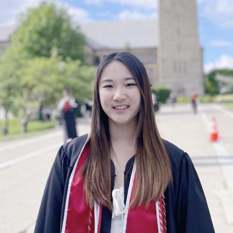 Joelle is standing in the middle of the frame. She is wearing a red stole over graduation regalia and a white dress. Behind her is a building that has been blurred out in the distance.