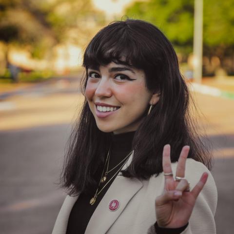 Jasmine is in the center of the frame, is facing slightly to her right. She is making a peace sign in the photo. She is wearing a tan blazer and a black turtle neck.