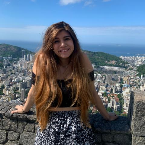 Paola is standing in the middle of the frame, leaned against a low stone railing and smiling at the camera. Behind her is a view of a city, rolling hills, and the ocean in the distance. 
