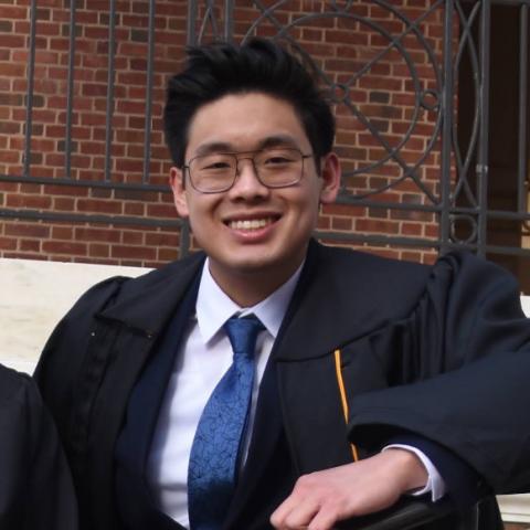 Trevor is standing in the middle of the frame, smiling at the camera. He is wearing square rimmed glasses. He is also wearing black graduation regalia over a navy suit with a white collared shirt and blue tie.