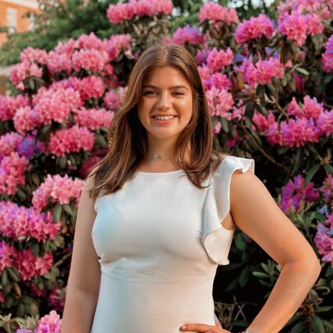 Emily Whalen is standing in the center of the frame, smiling at the camera. Her left arm is placed on her hip. Behind her is a bush of pink flowers that makes up the background of the photo.