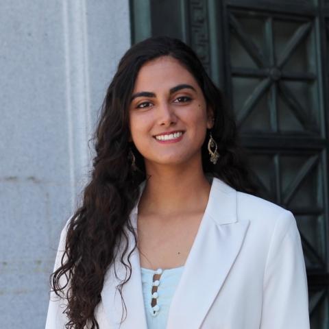 Alya is standing in the middle of the frame, facing the camera and smiling. She is wearing a white shirt under a white blazer.