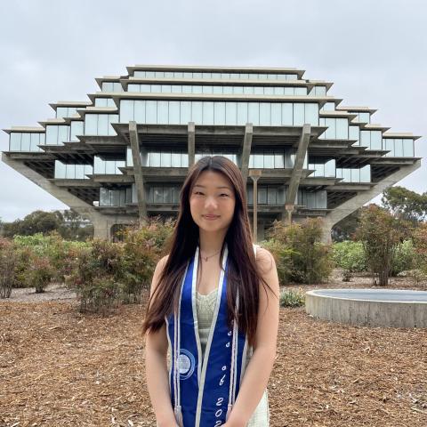 Sian Hong is standing in the middle frame, her arms crossed in front of her. She is wearing a graduation stole. Behind her is a beautiful ornate building in the background.