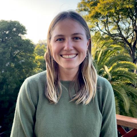 Morgan Prinz is standing in the center of the frame, facing the camera and smiling. Behind her is tree foliage and a setting sun.
