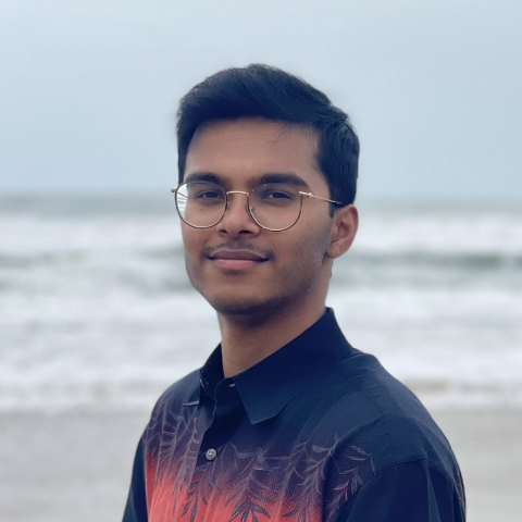 Shreyas is standing in the center of the frame, facing the camera and smiling. Behind him is the ocean with an overcast sky..