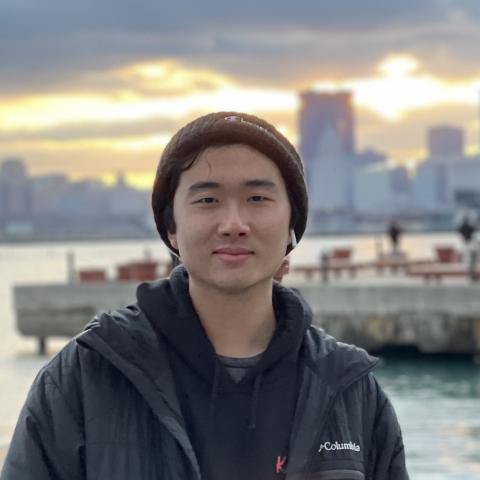 CJ is standing in the middle of the frame, facing the camera and smizing. In the background, there is a pier, sunset, and a city's skyline silhouette in the background.