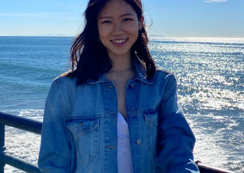 NHC SF AmeriCorps Member Clara Chung standing with a background at a pier. Ocean waves and shimmering sunlight frame her.