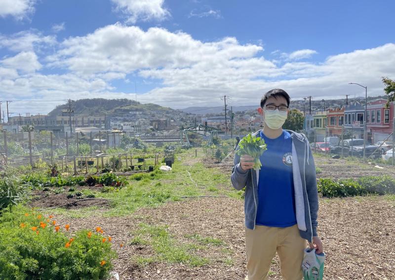 NHC SF member Jacky Chu holding a plant in their right hand. The background is a garden with semi-cloudy blue skies overhead. 