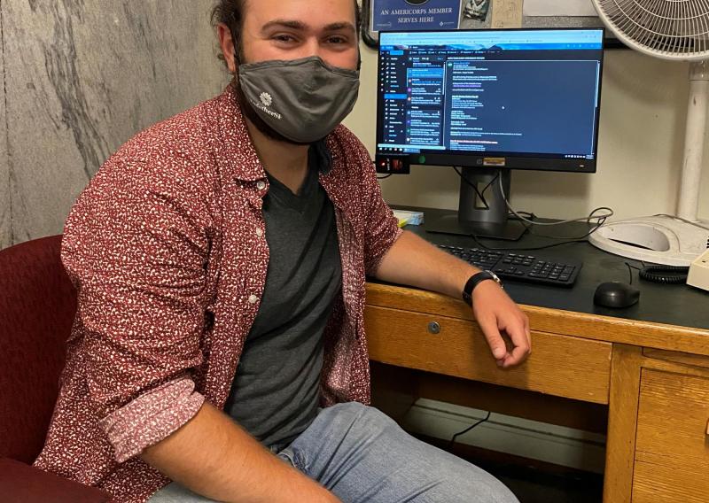 Eli Solomon slightly turned to the right, facing the camera. Eli is wearing a face covering, an open button down shirt with a grey undershirt, and lightwash jeans. He is sitting facing away from the computer and desk. 