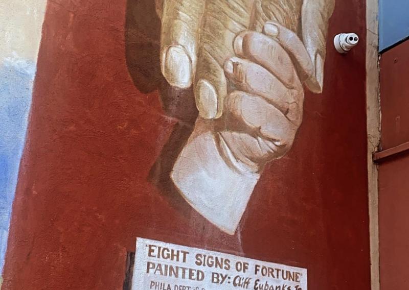 Mural of an adult hand being grasped by a childs hand on a red wall. Under the image is text, some illegible, but the last line is a quote that reads "my humanity is bound up in yours, for we can only be human together" - Desmond Tutu