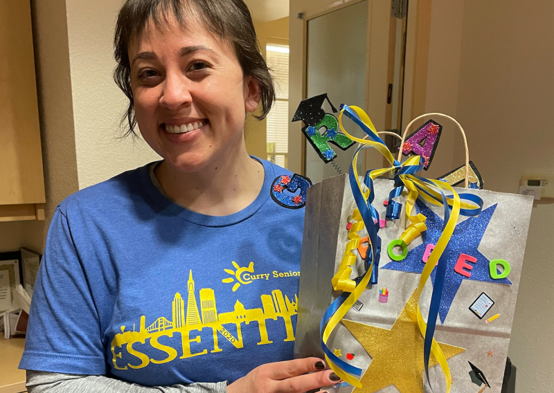 Hannah Griggs is seen here smiling towards the camera. She is holding a gift bag with ribbons of blue and yellow colors coming over the front of the bag. There is a yellow star on the bottom left front facing corner of the bag, and an adornment with a black graduation cap protruding from the top of the bag. 