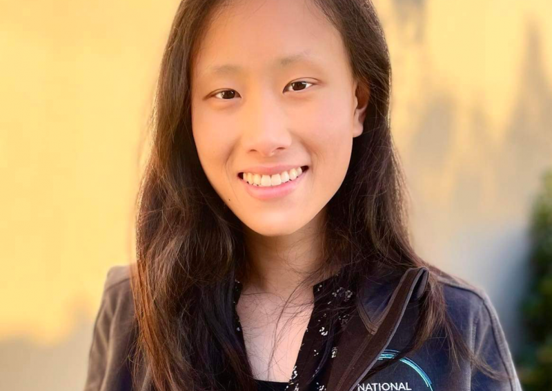 Mindy is facing the camera smiling, wearing a charcoal grey National Health Corps jacket over a blouse. Behind her is a cream-colored, blank wall.