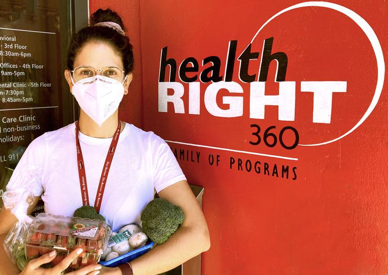 Mariana Carranza is seen standing in front of a HealthRIGHT 360 sign. She is holding assorted produce to give to patients. 