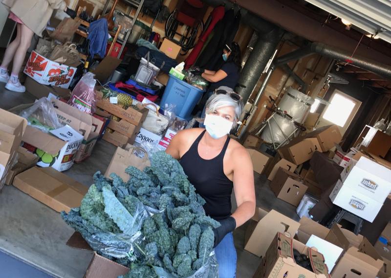 Janna is seen in the center of the frame, facing the camera with a mask on. She is squatting behind a box that contains kale, a green leafy vegetable to hand out to patients.