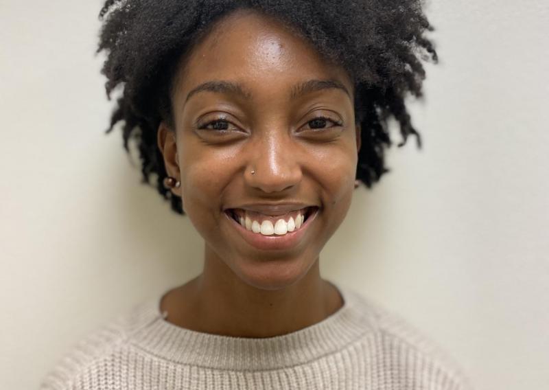 A person (Imani) is in the center of the frame smiling at the camera. She is wearing a knitted sweater. She is standing against a blank background.