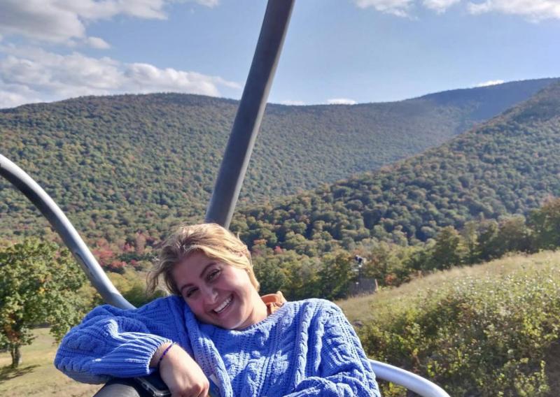 Sarah wearing a blue sweater sitting on a ski-lift during the summer, there are hills covered in green trees behind her.