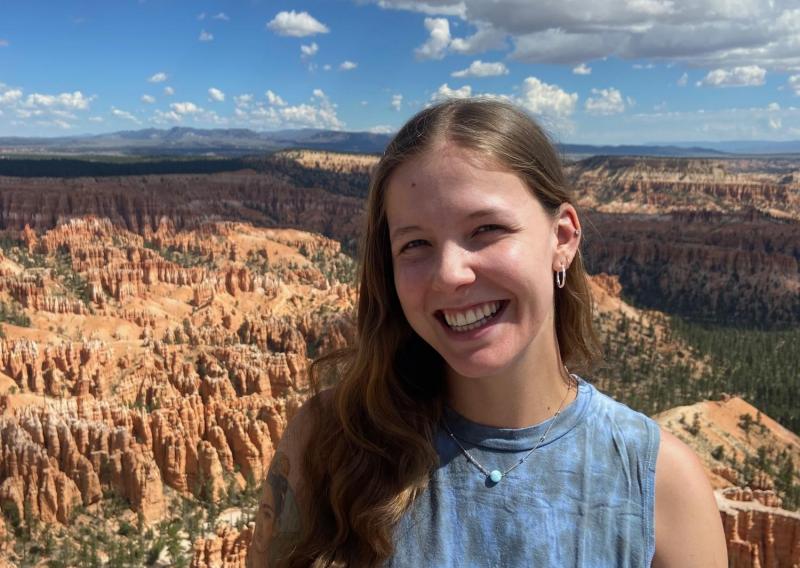 Grace Lemke is seen standing, facing the camera, smiling while wearing a tie-dyed tank top. 