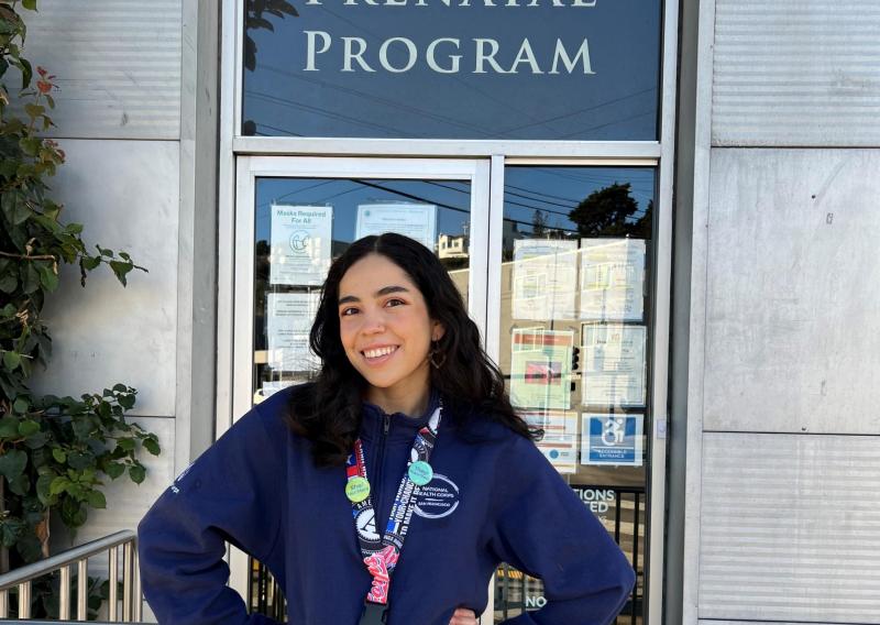 Jasmine is standing in the middle of the frame in front of the entrance to a building. Behind her and above the door is a sign that reads Homeless Prenatal Program. 