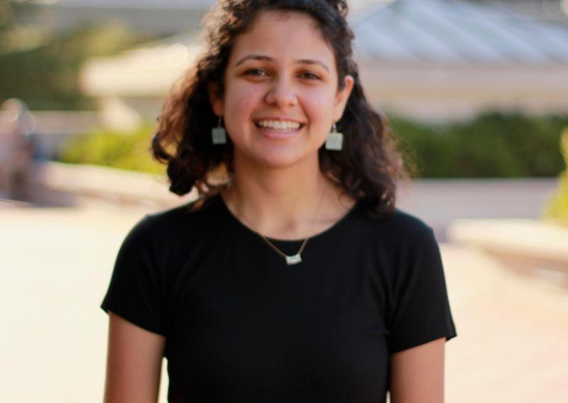 Saba Moussavian is standing in the center of the frame, facing the camera and smiling. 