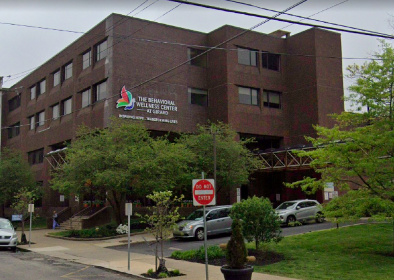 A brown brick building that says "The Behavioral Wellness Center at Girard" with green trees and some parked cars in the foreground