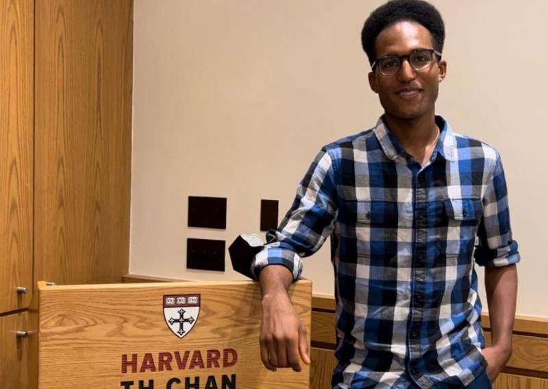 Spencer stands to the right of the frame, smiling at the camera. He is standing next to the podium showing the words "Harvard T.H. Chan School of Public Health"