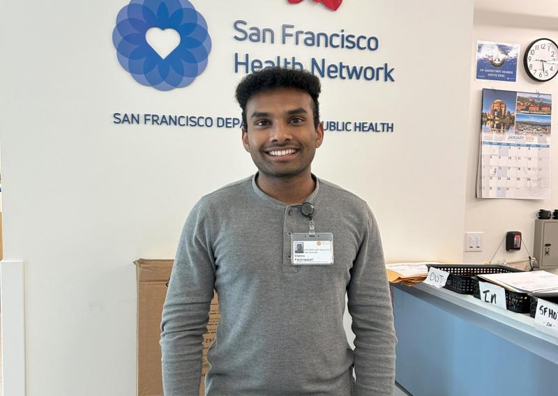Vishnu is standing in the middle of the frame, facing the camera. He is standing behind a podium and has his laptop set on the podium in front of him. Behind him is the San Francisco Health Network logo. 