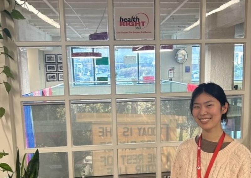 Hannah Min is facing the camera and smiling. She is standing in front of the check-in area for HealthRIGHT 360. 