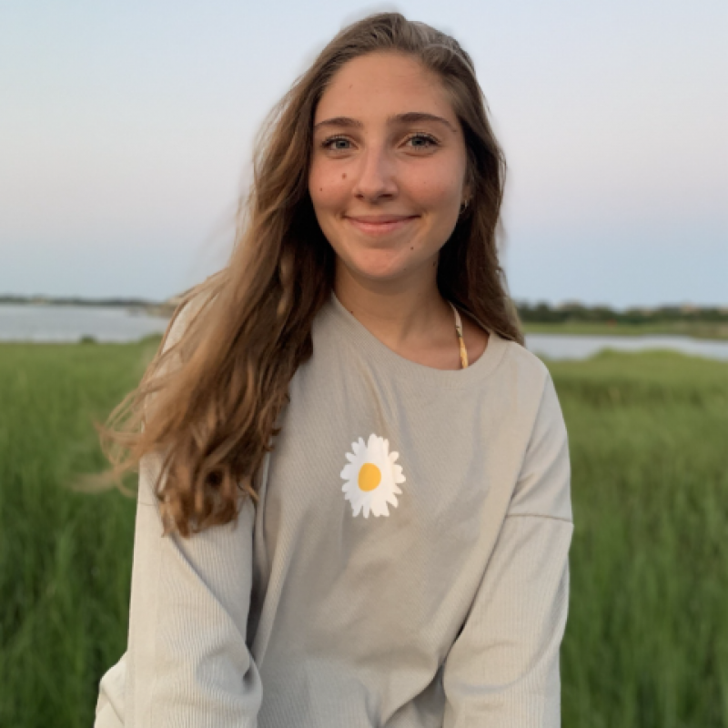 NHC PGH member Miranda soft-smiling in a field with a daisy on her sweatshirt