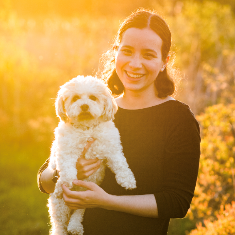 Aurora is standing facing the camera, smiling while carrying her dog, who is also looking at the camera for the photo. They are backlit and bathed in golden sunlight, with sunflower bushes to the left and a field to the right.