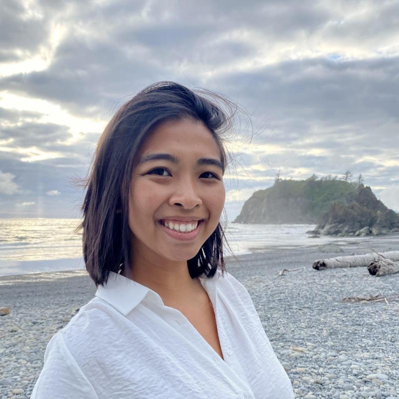 Emily Pham, facing the camera and smiling. Emily is wearing a white blouse. The sky is cloudy with some rays of sunlight breaking through over the grey-ish sand, ocean. There is an island in the distance