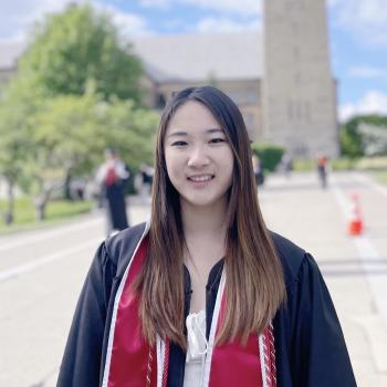 Joelle is standing in the middle of the frame. She is wearing a red stole over graduation regalia and a white dress. Behind her is a building that has been blurred out in the distance.