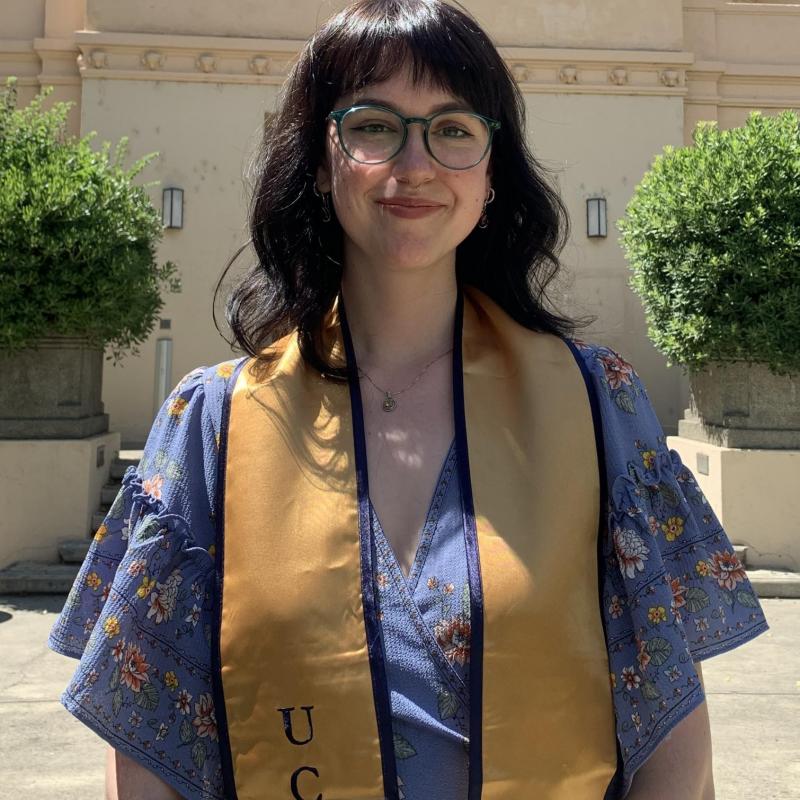 Sarah is standing in the middle of the frame, facing the camera. She is wearing a graduation stole over a lavender floral blouse. Behind her is a building with the letters "George Hart Hall" welded onto the face of the building. 