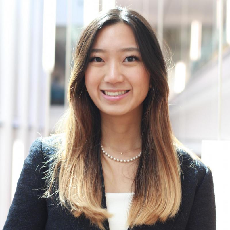 Sirapa is standing in the middle of the frame, facing the camera and smiling. She is wearing a charcoal blazer over a white blouse, with a pearl necklace around her neck.