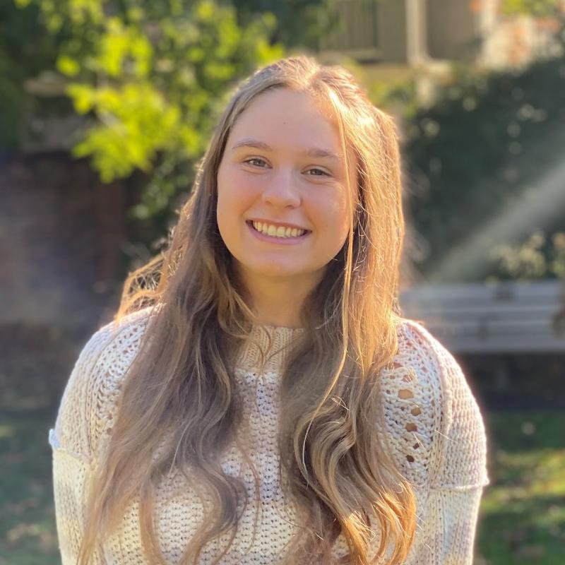 Hannah is standing in the middle of the frame, smiling at the camera, her hair laid over her shoulders. She is wearing a white sweater.