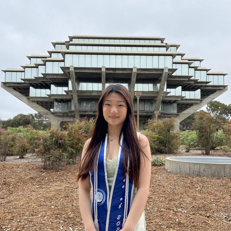 Sian Hong is standing in the middle frame, her arms crossed in front of her. She is wearing a graduation stole. Behind her is a beautiful ornate building in the background. 