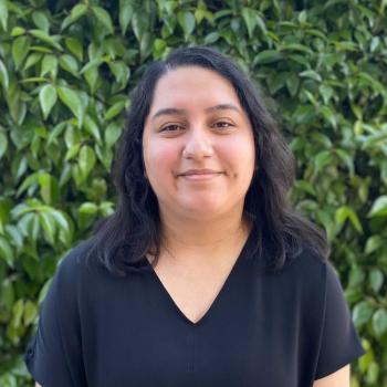 Melanie Lopez is standing in the middle of the frame, facing the camera and smiling. Behind her is a wall of leaves.