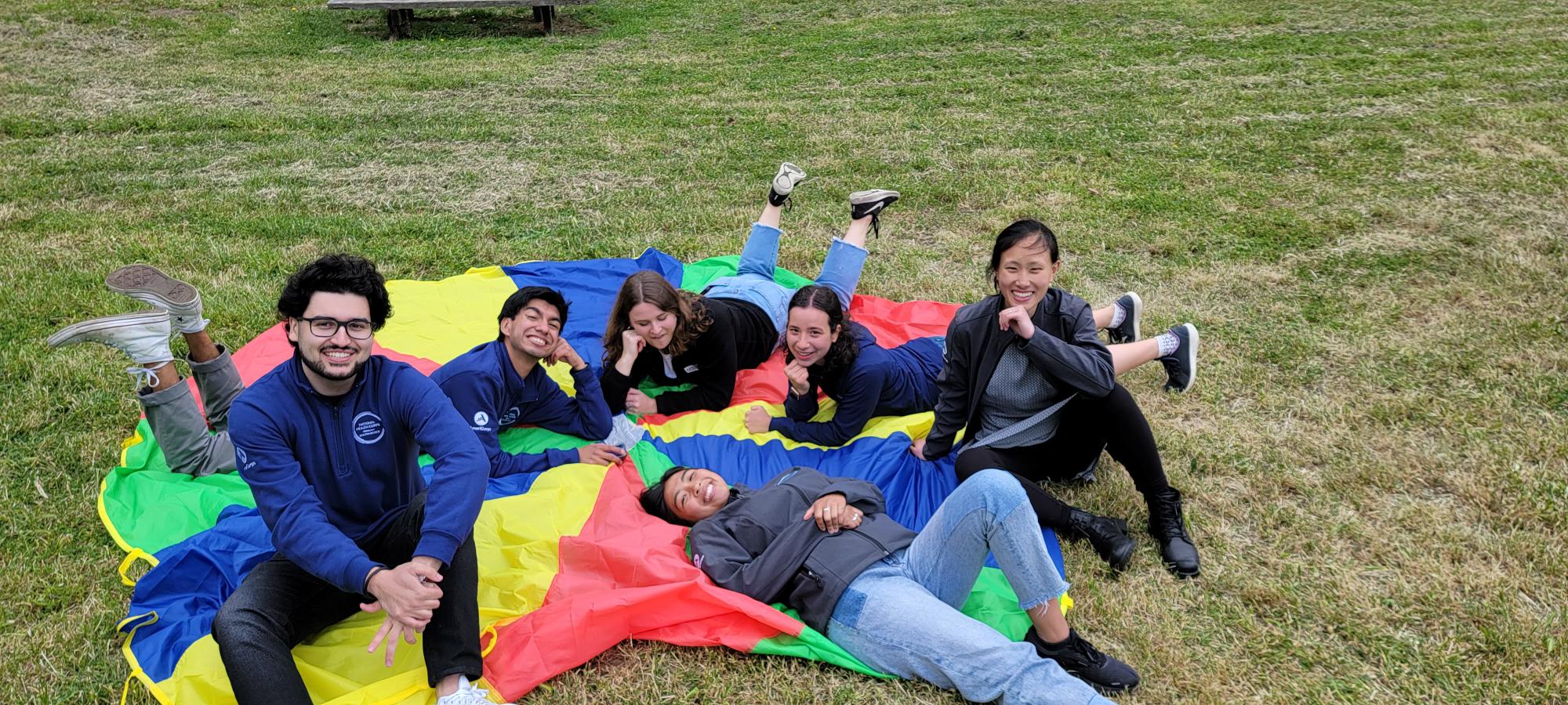 A group of Americorps members is seen sitting and laying on a rainbow cloth. From left to right: Jack Dokhanchi, Johnny Reyes, Autumn Kleinman, Emily Pham, Aurora Golden-Appleton, Mindy Dai.