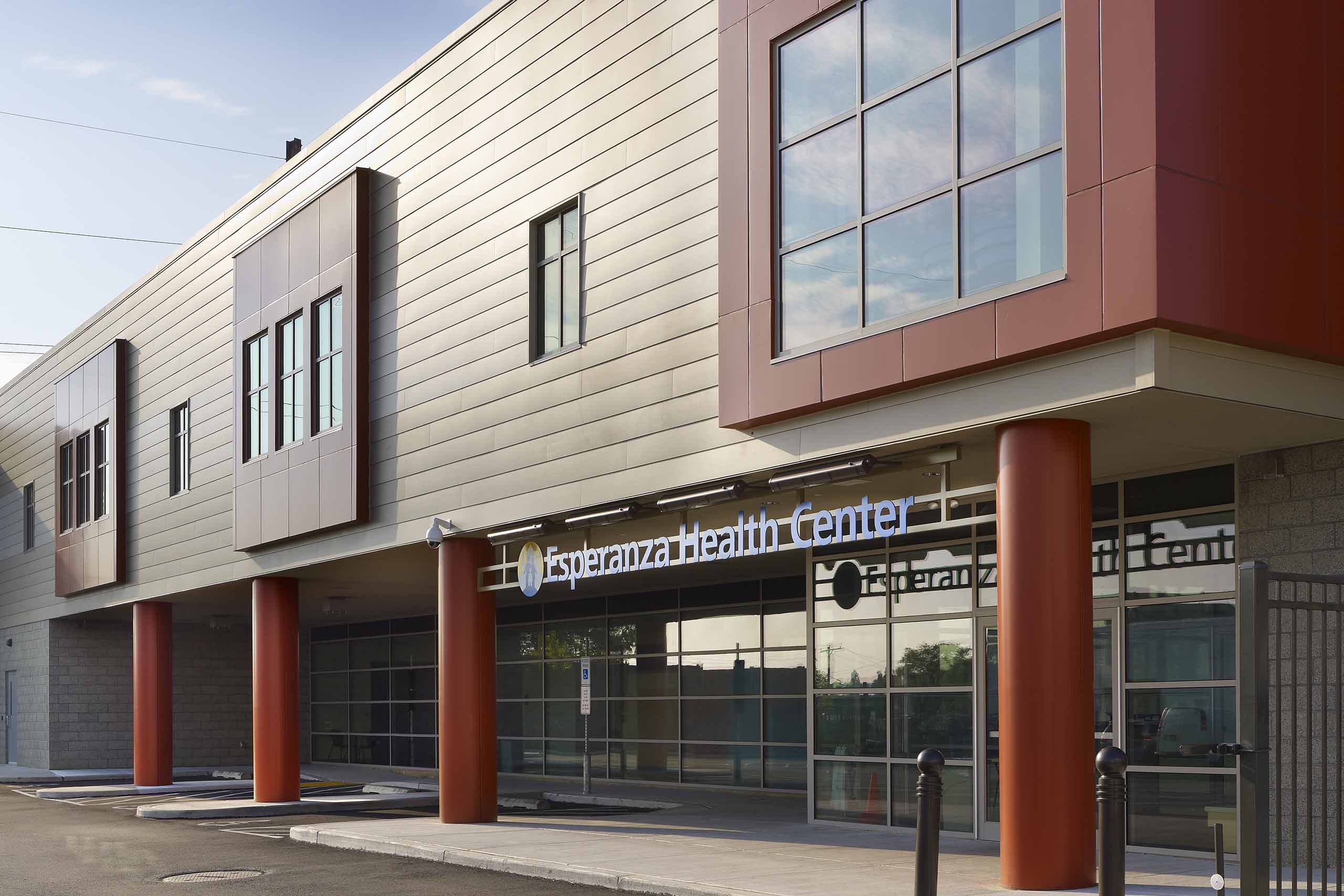 A two story building with tan siding and red columns. The first floor is mostly windows. There are three parking spots in the foreground. Above the entry to the building there is a sign that says "Esperanza Health Center"