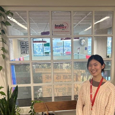 Hannah Min is facing the camera and smiling. She is standing in front of the check-in area for HealthRIGHT 360. 