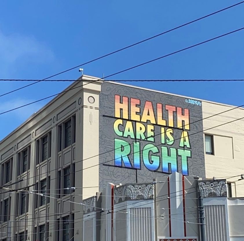A mural is seen on the side of a building stating "Health Care is a Right" in tall, bold font. The letters are colored in the array of a rainbow (red, orange, yellow, green, blue, purple).