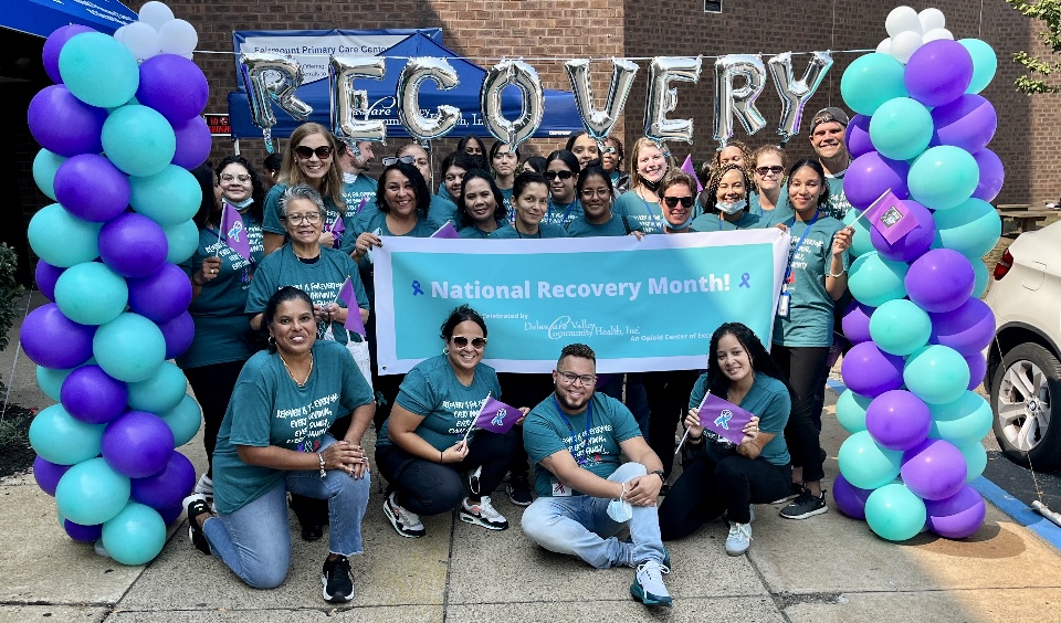 A group of people in blue shirts holding a banner that says "National Recovery Month." The group is standing between two pillars of balloons and below balloons that spell out the word recovery. Some members of the group are waving purple flags.